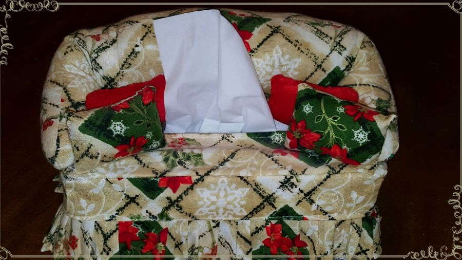 Image of a handmade miniature couch that is actually a tissue box cover. This one is a cream design with green, red, and gold Christmas designs in the material. It has miniature pillows in red and in matching material to the couch. There is one tissue popped up for retrieval. 