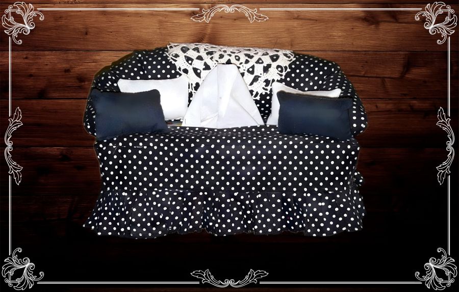 Image of a handmade miniature couch that is actually a tissue box cover. It's black with white polka dots, black and white miniature pillows, and a white lace doily over the back. One tissue is popped up and ready for retrieval. The background has been replaced by wood grain with an artistic white frame. 