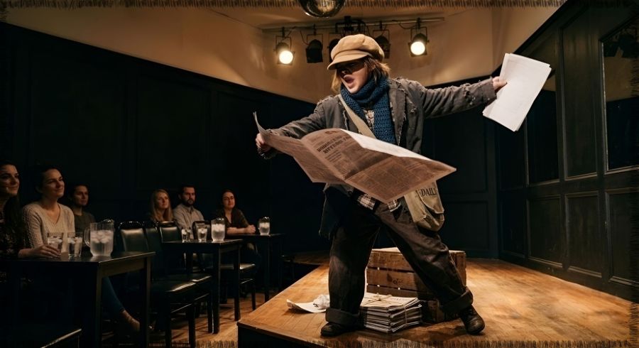 An animated theater stage recreation from the 1980s show "How the West Was Lost" at Aquarius Rising. A young performer under a warm spotlight wears a tan newsboy cap, a tattered gray jacket over a plaid shirt, a blue crocheted scarf, and baggy dark brown cuffed pants dragging on the hardwood floor. The performer dynamically leans forward, arms flung wide while holding an open, full-size newspaper as if dramatically shouting a headline. A tied stack of papers sits on the stage at their feet. In the dark background audience area, small black wooden tables hold clear glasses of ice water. Image and alt text by Gemini from Google. 